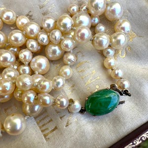 May include: A vintage pearl necklace with a green jade clasp. The necklace features multiple strands of cream-coloured pearls of varying sizes. The jade clasp is oval-shaped and a vibrant green colour, contrasting the pearls.