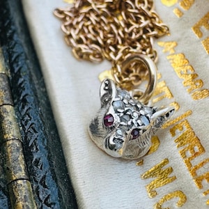 Puede incluir: Collar con colgante de plata con cabeza de gato, ojos de rubí y cara con diamantes. El colgante cuelga de una cadena dorada. El collar se muestra sobre una superficie blanca.