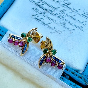 May include: A pair of gold dragonfly stud earrings. Each earring features a gold frame with ruby, sapphire, emerald, and diamond accents. The earrings are displayed in a blue and white box with handwritten text.