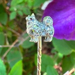 May include: A silver-coloured dog-shaped pin adorned with sparkling blue and green gemstones. The pin features a twisted gold-coloured stem. The background is blurred with green foliage and a purple flower.