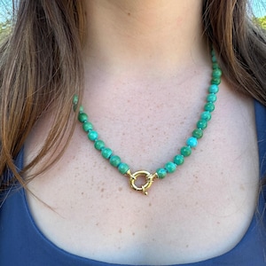 May include: A beaded necklace featuring round, variegated turquoise-colored stones in shades of green and blue. The beads are strung together and secured at the front with a prominent, circular gold-toned metal spring ring clasp closure.