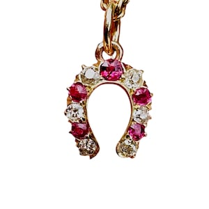 Könnte beinhalten: Ein goldener Hufeisenanhänger mit abwechselnd rund geschliffenen roten Rubinen und klaren weißen Diamanten. Der Anhänger hängt an einem kleinen goldenen Verbindungsring und einer zarten Goldkette und zeigt eine symmetrische Anordnung der Edelsteine.