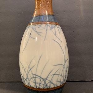 May include: A ceramic vase with a white background and blue and brown accents. The vase has a narrow neck and a wide, rounded body. The design features a pattern of thin, blue lines that resemble grass.