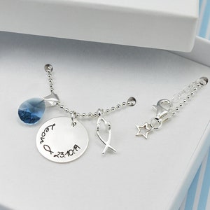 May include: A silver necklace with a blue crystal pendant, a round charm with text, and a fish charm, presented in a white box. The box has a decorative scalloped edge with blue accents. The text on the charm reads "Leon" and "23.10.19".