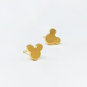 May include: A pair of gold-colored stud earrings in the shape of Mickey Mouse's head. The earrings are small and simple, with a post backing. The earrings are set against a white background.