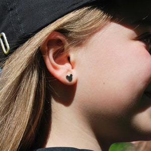 May include: Black Mickey Mouse-shaped stud earrings. The earrings are small and sit on the earlobe. The person is wearing a black baseball cap and a black shirt. The background is blurred.