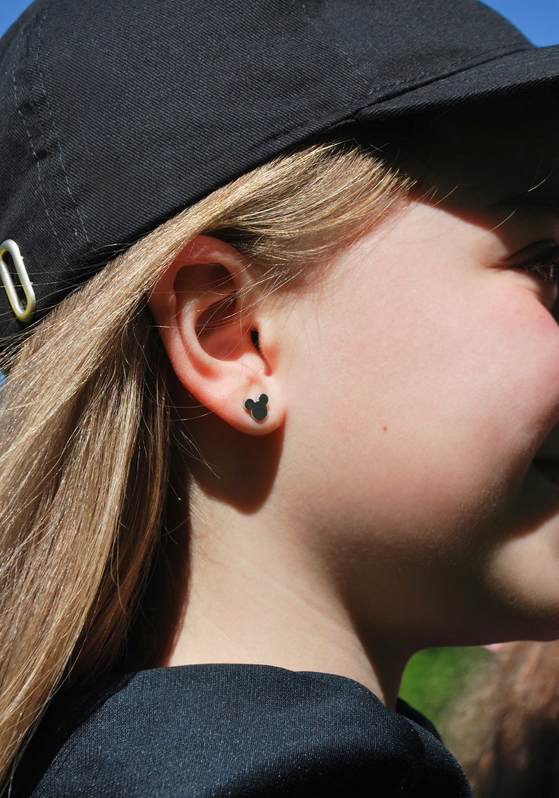 May include: Black Mickey Mouse-shaped stud earrings. The earrings are small and sit on the earlobe. The person is wearing a black baseball cap and a black shirt. The background is blurred.