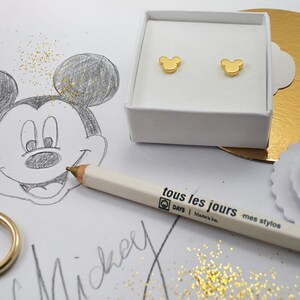 May include: A pair of gold-colored Mickey Mouse stud earrings displayed in a white box. A pencil with the text "tous les jours" and a sketch of Mickey Mouse are also visible.