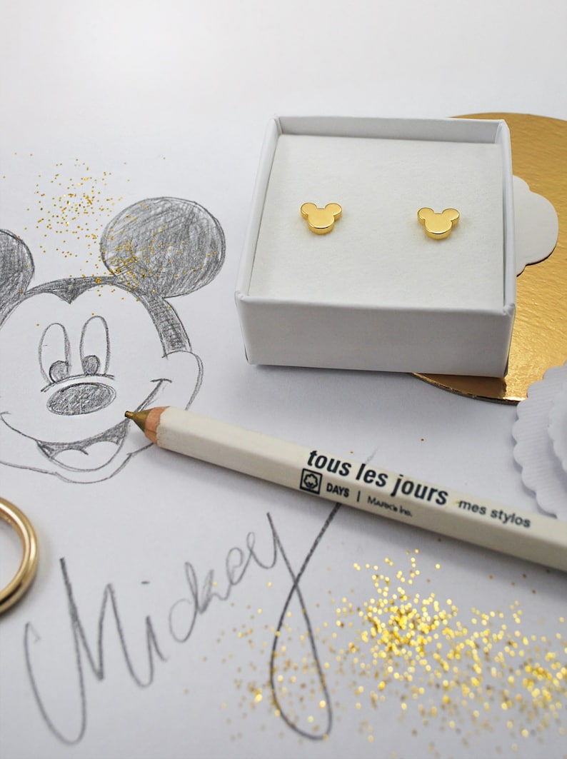May include: A pair of gold-colored Mickey Mouse stud earrings displayed in a white box. A pencil with the text "tous les jours" and a sketch of Mickey Mouse are also visible.