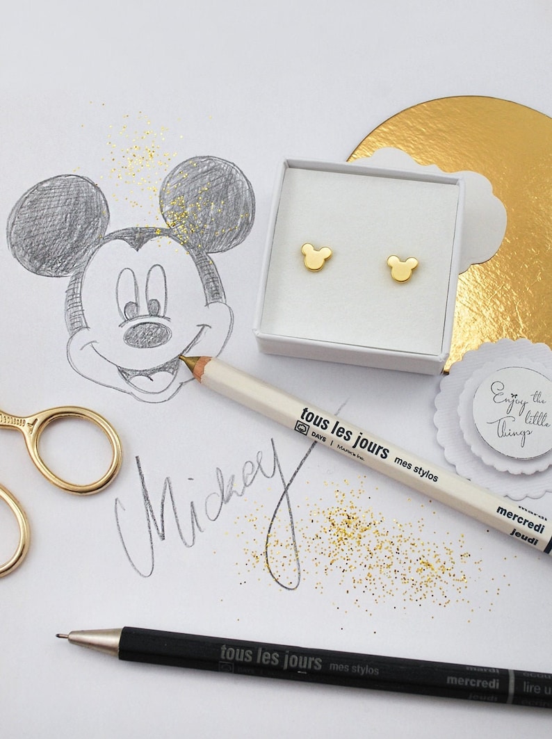 May include: Gold-colored Mickey Mouse-shaped earrings in a white box, next to a pencil sketch of Mickey Mouse. The word "Mickey" is handwritten in cursive. A white pencil with the text "tous les jours mes stylos" is also present.