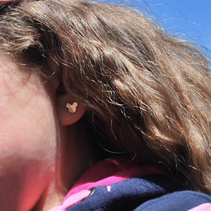 May include: A close-up of a person's ear with a gold-colored Mickey Mouse-shaped stud earring. The person is wearing a pink hat and a navy blue and pink hooded sweatshirt. The background is a clear blue sky.