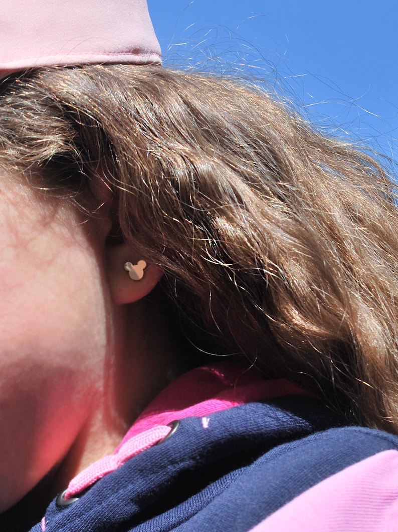 May include: A close-up of a person's ear with a gold-colored Mickey Mouse-shaped stud earring. The person is wearing a pink hat and a navy blue and pink hooded sweatshirt. The background is a clear blue sky.