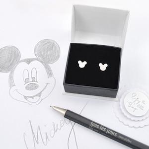 May include: Silver Mickey Mouse stud earrings presented in a black box with a white interior. A pencil sketch of Mickey Mouse and the word "Mickey" are visible. A pen with the text "tous les jours" is also present.
