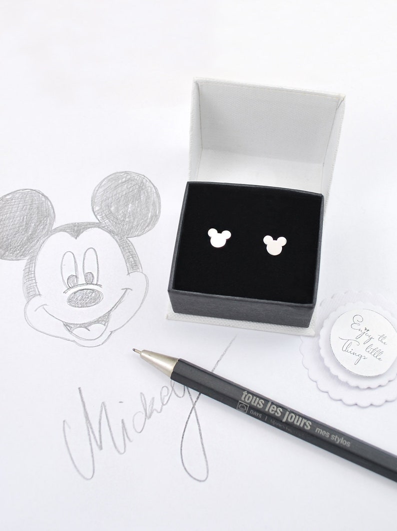 May include: Silver Mickey Mouse stud earrings presented in a black box with a white interior. A pencil sketch of Mickey Mouse and the word "Mickey" are visible. A pen with the text "tous les jours" is also present.