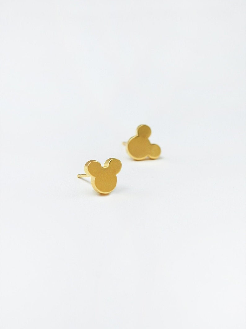 May include: A pair of gold-colored stud earrings in the shape of Mickey Mouse's head. The earrings are small and simple, with a post backing. The earrings are set against a white background.
