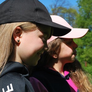 May include: Two baseball caps, one black and one pink, are worn by two people. The black cap is in the foreground, and the pink cap is behind it. The people are wearing black and pink hoodies. The background is a bright blue sky and green trees.