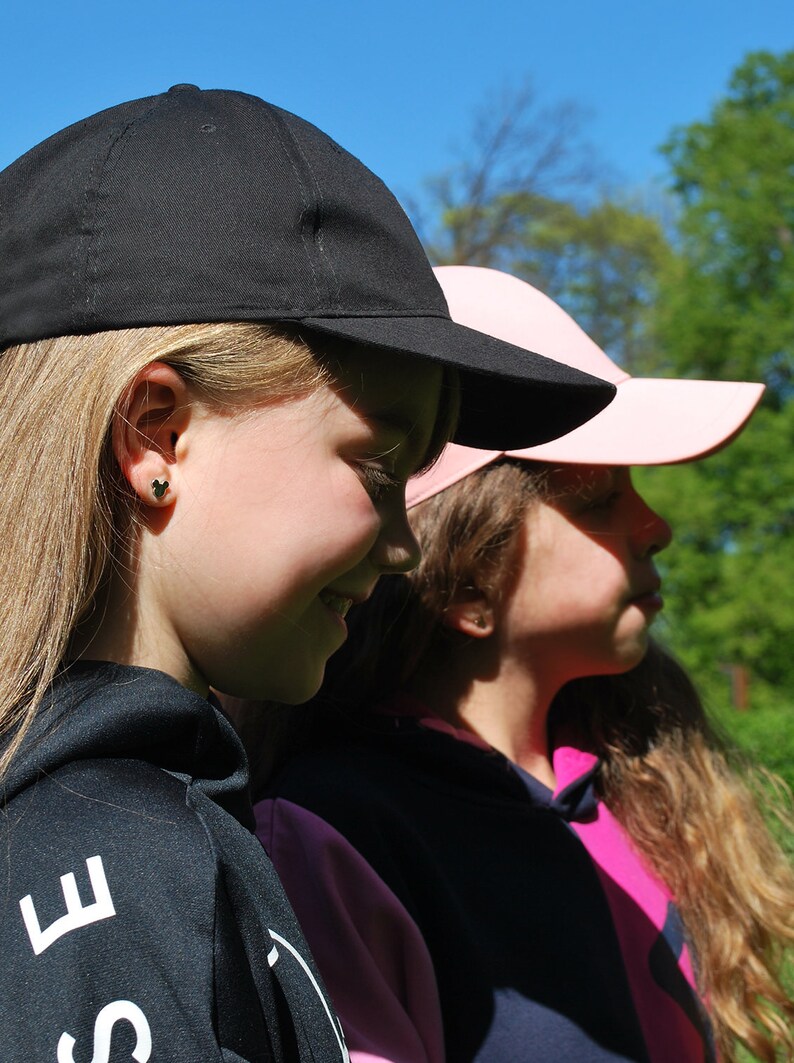 May include: Two baseball caps, one black and one pink, are worn by two people. The black cap is in the foreground, and the pink cap is behind it. The people are wearing black and pink hoodies. The background is a bright blue sky and green trees.