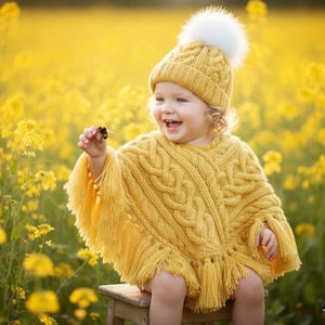 May include: A yellow knit poncho featuring a cable knit pattern and long fringe trim along the hem, paired with a matching yellow knit beanie topped with a large white pom-pom. The garments are crafted with a textured, chunky yarn design.