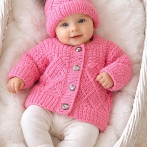 May include: A bright pink knitted cardigan with a textured diamond pattern and four buttons down the front, paired with a matching pink knitted beanie featuring a large, fluffy brown pom-pom on top and plain white leggings.