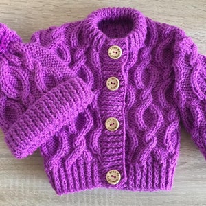 Peut inclure: Un ensemble composé d'un cardigan et d'un bonnet tricotés à la main en violet vif. Le cardigan présente un motif en torsades, une encolure ronde et quatre boutons en bois clair portant l'inscription Aran By Anderson sur chaque bouton.