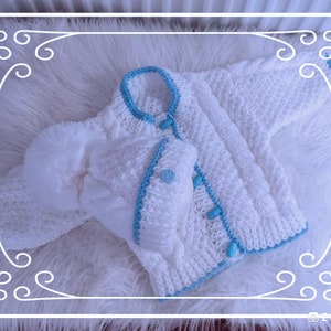 May include: A white knitted baby jacket and matching hat with a fluffy pom-pom. The set features light blue trim and buttons. The jacket has long sleeves and a button-up front. The set is displayed on a white, fluffy surface.