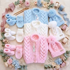 May include: A collection of three baby sets in light blue, white, and soft pink. Each set includes a cable-knit cardigan with buttons, a matching beanie with a large pom-pom, booties, and mittens. The items are arranged with floral accents.