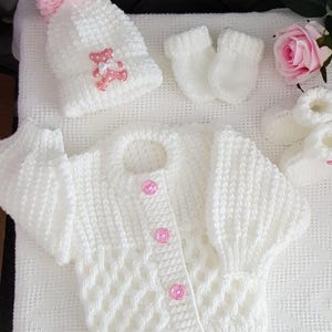 May include: A white knitted baby set consisting of a long-sleeved cardigan with four pink buttons, a matching beanie hat with a pink pom-pom and a pink bear appliqué, a pair of white mittens, and a pair of white booties with pink accents.