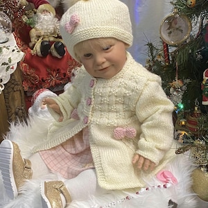 Puede incluir: Una muñeca vestida con un conjunto de punto color crema, que incluye un gorro con pompón y una rebeca adornada con lazos rosas. La muñeca lleva un vestido de cuadros rosas, medias blancas y zapatos dorados con purpurina. Escena navideña.