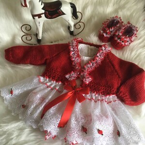 May include: A red knitted cardigan with white lace trim and a matching white lace skirt with red ribbon bow. The skirt has embroidered red roses.
