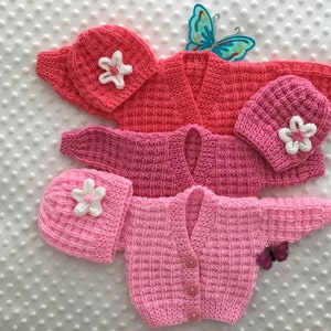 Puede incluir: Dos conjuntos de ropa de bebé de punto, uno rosa y otro coral, cada uno con un gorro a juego. Los gorros tienen una aplicación de flor blanca. Los jerséis son cárdigans con cierre de botones.