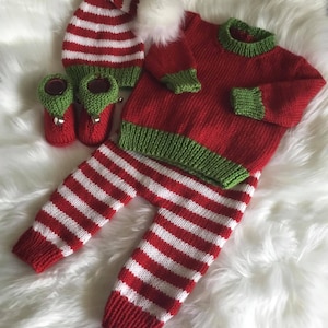 Könnte beinhalten: Ein gestricktes Baby-Outfit in Rot und Weiß mit Streifen, bestehend aus Mütze, Pullover und Hose. Der Pullover hat grüne Borten und die Mütze einen weißen Bommel.