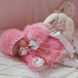 Op de afbeelding: Een roze gebreide baby-outfit met een bijpassende muts en slofjes. De outfit is gemaakt van zacht, pluche garen en heeft een knoopsluiting. De baby draagt een speen.