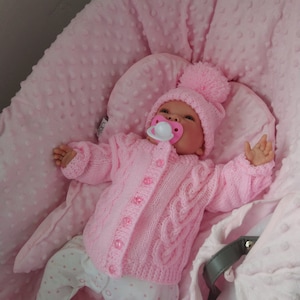 May include: A pink knitted baby doll outfit with a white dummy, a pink knitted hat with a white pom-pom, and white trousers with pink polka dots. The doll is lying in a pink and white padded baby seat.