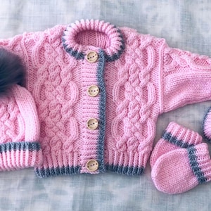 May include: A pink knitted baby cardigan, hat and booties set with grey trim. The cardigan has a cable knit pattern and buttons down the front. The hat has a grey pom-pom on top.