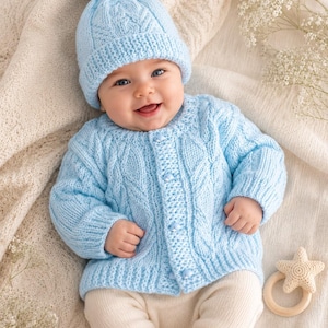 May include: A light blue cable-knit cardigan with matching buttons and a coordinating beanie featuring a pom-pom on top. The set is paired with cream-colored leggings and a small, beige, star-shaped crochet rattle with a wooden ring handle.