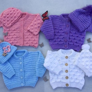Könnte beinhalten: Vier gestrickte Baby-Cardigans in Rosa, Lila, Blau und Weiß mit passenden Mützen. Jeder Cardigan hat einen Knopfverschluss und ein strukturiertes Strickmuster.