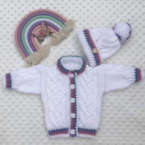 May include: A white cable-knit baby cardigan, hat, and rainbow wall hanging. The cardigan and hat have colourful trim and button closures. The rainbow has pastel colours and a butterfly accent. A charming set for a baby.