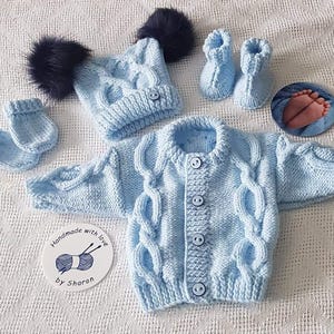 May include: A light blue knitted baby cardigan with a cable knit pattern and buttons. The set also includes a matching hat with pom poms and booties.
