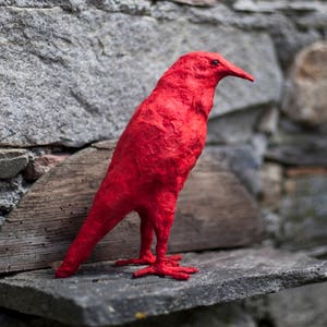 May include: A bright red paper mache bird sculpture perched on a stone ledge. The bird has a long beak and is facing to the right. The background is a stone wall with a green plant growing on the right side.