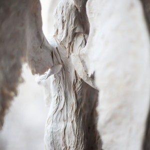 May include: Two white paper mache angels with wings embracing each other. The angels are made of textured paper and have a rustic, handmade look.