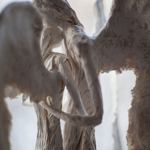 May include: A close-up of a white paper mache sculpture of three angels with wings. The angels are intertwined and appear to be embracing.