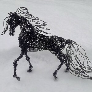 Puede incluir: Una escultura de alambre negro de un caballo de pie sobre una superficie nevada. La crin y la cola del caballo son largas y fluidas, y sus patas est&aacute;n dobladas como si estuviera en movimiento.