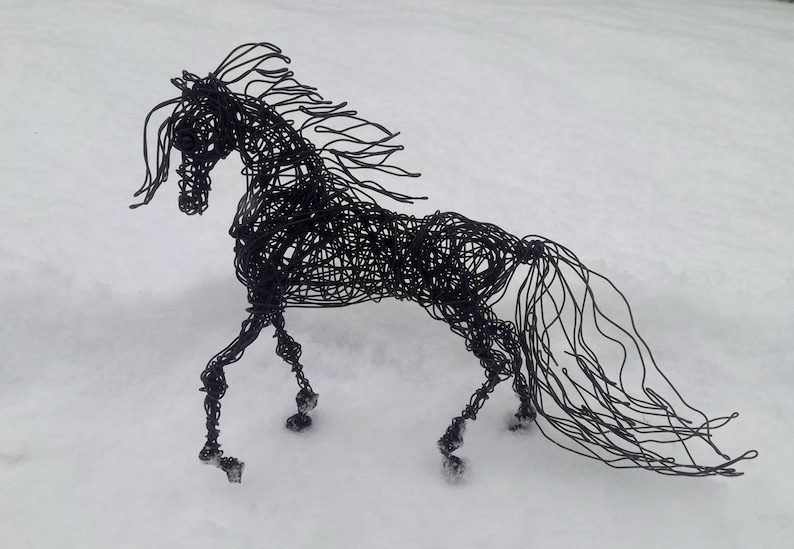 Puede incluir: Una escultura de alambre negro de un caballo de pie sobre una superficie nevada. La crin y la cola del caballo son largas y fluidas, y sus patas est&aacute;n dobladas como si estuviera en movimiento.