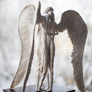May include: A white paper mache sculpture of two angels with wings, standing back to back, with one angel embracing the other.