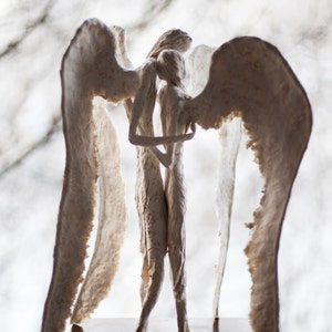 May include: A white sculpture of two angels with wings embracing each other. The sculpture is on a wooden base.