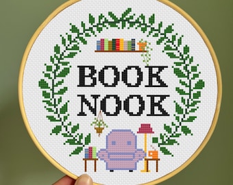 May include: A cross-stitch embroidery hoop with a green leafy wreath surrounding the text "BOOK NOOK". Inside the wreath is a purple armchair, a small table with books, a lamp, and a cup of tea.