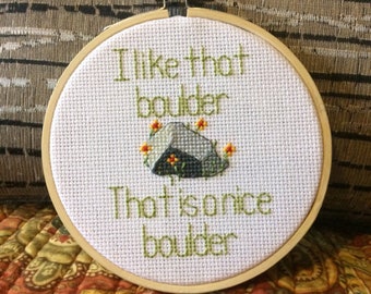 May include: A white embroidery hoop with a green cross-stitch design that reads "I like that boulder. That is a nice boulder."  A small gray rock with orange flowers is stitched in the center of the hoop.