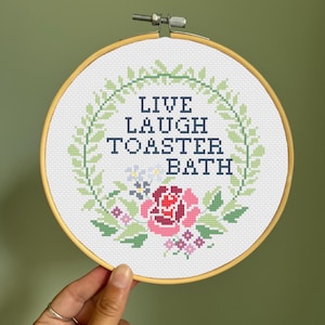 Peut inclure: Un cercle de broderie au point de croix avec l'inscription "LIVE LAUGH TOASTER BATH" en bleu marine. Le motif comprend une couronne florale avec des feuilles vertes, des roses roses et rouges, et de petites fleurs blanches et violettes. Le cercle est tenu par une main.