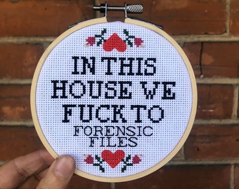 May include: A white embroidery hoop with the text "In this house we f*ck to forensic files" in black lettering. The text is surrounded by red hearts and green leaves.