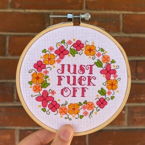 May include: A round embroidery hoop with a white fabric background. The fabric is embroidered with a wreath of pink and orange flowers and green leaves. The text "JUST FUCK OFF" is embroidered in pink letters in the center of the wreath.